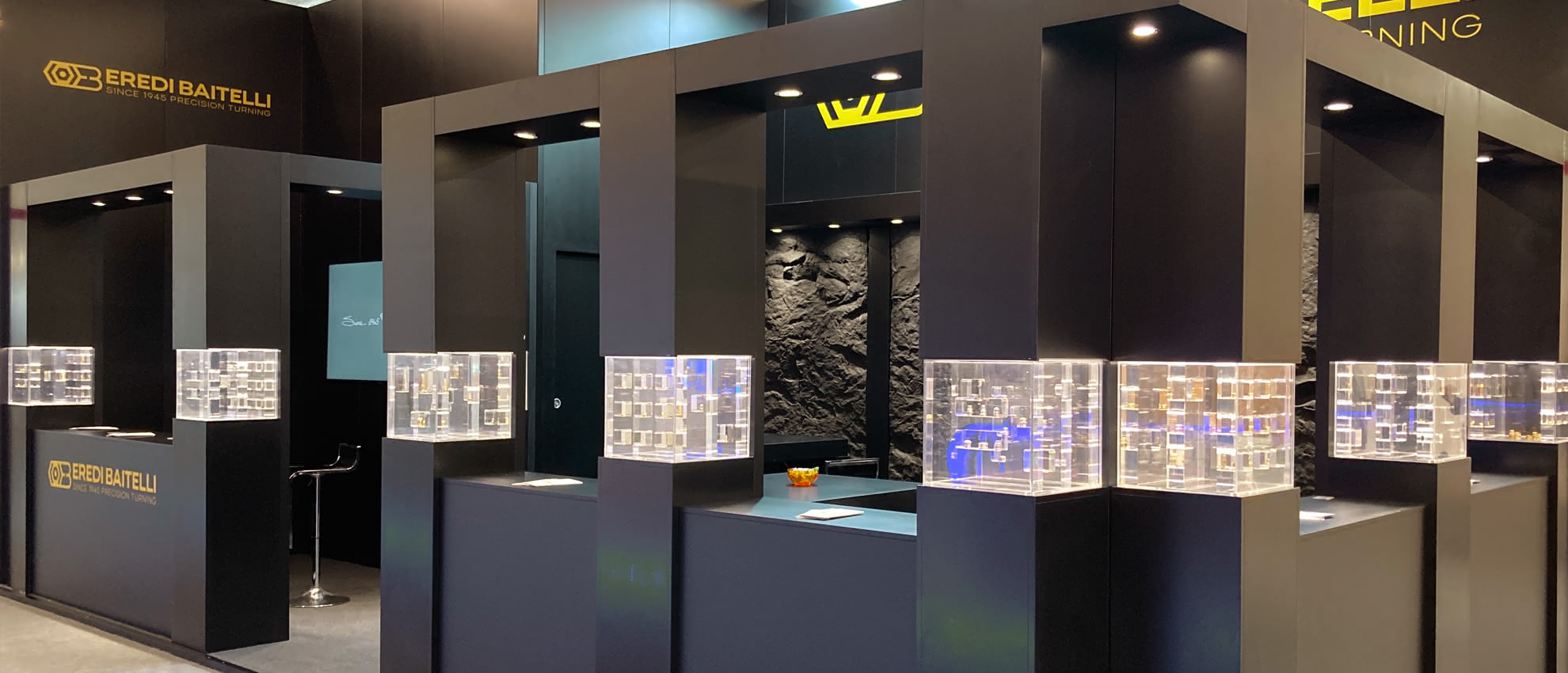 Vue latérale du stand Eredi Baitelli lors d’un salon professionnel international, avec structures modulaires noires et vitrines transparentes disposées en îlot.
